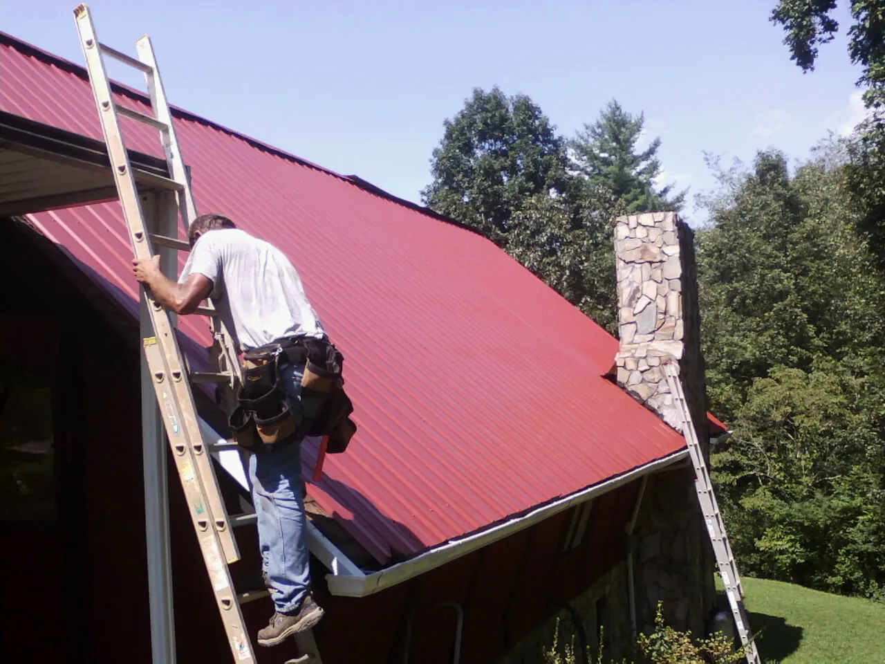 Roofer on a ladder beside a red metal roof and stone chimney — Soffit & Fascia Repair by Roofing Brothers in Fombell